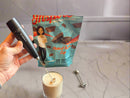 A hand holds a black frother above a glass of blended protein powder, while a colorful bag of peanut butter cup protein powder is visible in the background on a marble countertop.