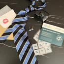 A black and blue striped tie is laid on a dark surface alongside a small black bottle, cufflinks, a card with a kiss mark, and a package of male libido patches labeled "Ignite Him."