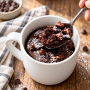 A hand holds a spoonful of chocolate mug cake topped with chocolate chips and powdered sugar, resting in a white mug on a wooden table, with a checkered cloth and chocolate chips in the background.