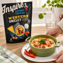 A hand reaches for a green dish of a Western omelet topped with vegetables and sausage, with a packet of Inspire protein kitchen Western omelet mix displayed in the background on a wooden table.