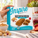 A hand holds a blue box of Inspire Peanut Butter & Jelly protein bars, with a bowl of peanut butter, strawberries, and jelly nearby, set on a bright kitchen counter.