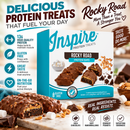 A box of Inspire Rocky Road protein bars is displayed with one bar broken open, showcasing its chocolate coating, soft marshmallows, and crunchy peanuts, against a backdrop of chocolate pieces and nuts.