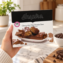 A hand holds a box of Inspire Choc-a-lot Chip Protein Bars, showcasing the product alongside chocolate chips and pieces on a kitchen counter with a bright, inviting background.