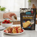 A hand pours syrup from a small pitcher onto a plate of silver dollar pancakes topped with strawberries, next to a bag of Inspire Protein Kitchen Silver Dollar Pancake Mix in a bright kitchen setting.