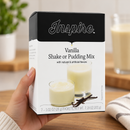 Hand holding a box of Inspire Vanilla Shake or Pudding Mix in a bright kitchen. Package features creamy vanilla shake and pudding with vanilla beans.