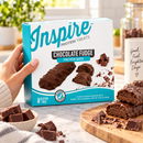 Hand holding a box of Inspire Chocolate Fudge Protein Bars on a bright kitchen counter, surrounded by chocolate pieces and chocolate-covered protein bars.