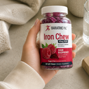 A hand holds a bottle of Bariatric Pal Iron Chew in sour raspberry flavor, featuring a label that highlights 45mg of iron, alongside a cozy setting with a glass of water and a soft blanket.