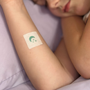 A child is resting with their eyes closed, showing a bandage on their arm featuring a green design, against a soft purple background.