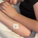 A child is resting with their arms crossed on a striped bed, featuring a decorative bandage with a moon and stars design on their forearm.
