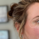 A woman with wavy hair and earrings looks to the side, showcasing a translucent skincare patch applied near her eye against a softly blurred background.