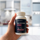 A hand holds a bottle of Bariatric Pal Multivitamin One, labeled for daily use with 45mg of iron, against a blurred kitchen background.