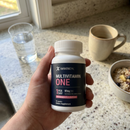A hand holds a white bottle of BariatricPal Multivitamin One in front of a kitchen counter with a glass of water, a mug, and a bowl of food.