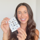 A woman with long, wavy hair smiles brightly while holding a pack of six decorative stickers featuring snowflake designs against a light background.
