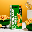A green box of Kaizen Protein Mac & Cheese with a classic cheddar flavor stands next to a bowl of creamy macaroni, set against a bright orange background, highlighting its nutritional benefits.