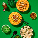 Three bowls of pasta with different sauces and toppings are arranged on a bright green surface, accompanied by a green drink, a small bowl of red pepper flakes, and chopped green onions.