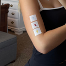 A person with light skin is showing their arm, which has three colorful adhesive bandages (blue, red, and yellow) on it, while sitting in a cozy room with a gray couch and white furniture in the background.