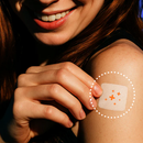 A smiling woman with long red hair gently touches a bandage on her upper arm, showcasing its design against a softly lit background.