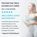 A woman in a light gray athletic outfit holds a skincare product while standing against a white brick wall, showcasing a positive review highlighting its moisturizing benefits and pleasant scent.
