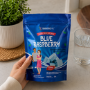 A hand holds a blue stand-up pouch of BariatricPal Blue Raspberry soft chew dietary supplement. The package is blue with white text and illustrations of a couple and blue raspberry pieces.