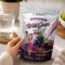 A hand holds a package of "Wild Grape" calcium citrate soft chews, featuring illustrations of a man and woman, with a glass of water and a bowl of fruit in a bright kitchen setting.