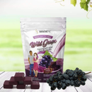 A stand-up pouch of BariatricPal Wild Grape flavored calcium citrate soft chew gummies, shown on a light wood surface with several dark purple gummies blocks in front and a bunch of dark grapes to the right. The background is a soft-focus green gradient.