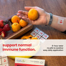 A hand holds an orange above a wooden tray with strawberries and oranges, while a patch with a cherry design is visible on the wrist, promoting immune support with a product label nearby.