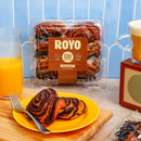 A yellow plate holds two chocolate rolls with a swirl pattern, next to a glass of orange juice and a cup of coffee, with a package of Royo chocolate rolls in the background against a light blue wall.