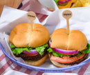 Two freshly made burgers with lettuce, tomato, and onion are placed in a blue basket, each topped with a wooden pick labeled "HERO," set against a red and white checkered tablecloth.