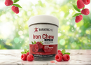 A container of Bariatric Pal Iron Chew, labeled "Sour Raspberry," sits on a light surface surrounded by fresh raspberries, with a blurred green background suggesting a natural setting.