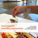 A hand with a patch on the wrist picks up a toothpick with an olive and ham, while a colorful salad and a platter of snacks are visible on a table in a bright, modern kitchen.