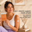 A woman in a sleeveless top smiles while holding a package of Iron Plus Patch, which is applied to her arm, in a softly lit indoor setting with decorative plants.