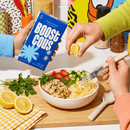 Two hands prepare a meal by squeezing lemon over a bowl of couscous topped with grilled chicken, cherry tomatoes, and cucumbers, while a colorful box of "Boost Cous" is held in the background.