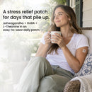 A woman with long hair sits comfortably on a porch, holding a cup and smiling, while wearing a stress relief patch on her arm, surrounded by plants and a cozy setting.