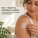 A woman gently places a daily patch on her upper arm, showcasing a product that contains plant-based iron and vitamin C, with a soft, natural background and greenery nearby.