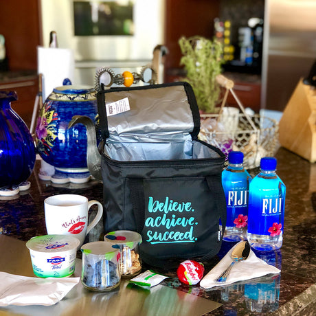A black lunch bag with the text "Believe. Achieve. Succeed." sits on a kitchen counter surrounded by snacks, two bottles of Fiji water, a mug, and various food containers.
