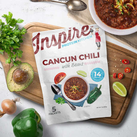 A package of Inspire Protein Kitchen Cancun Chili with Beans is displayed on a wooden cutting board, surrounded by fresh ingredients like avocado, bell pepper, and herbs, with a bowl of chili nearby.