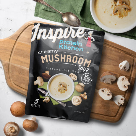 A packet of Inspire Creamy Mushroom Soup is displayed on a wooden cutting board alongside sliced mushrooms and a bowl of soup, with a spoon resting nearby on a green cloth.