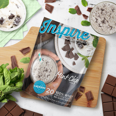 A hand holds a glass of mint chip protein shake with a straw, surrounded by chocolate pieces and fresh mint, with a package of Inspire protein powder prominently displayed on a wooden cutting board.